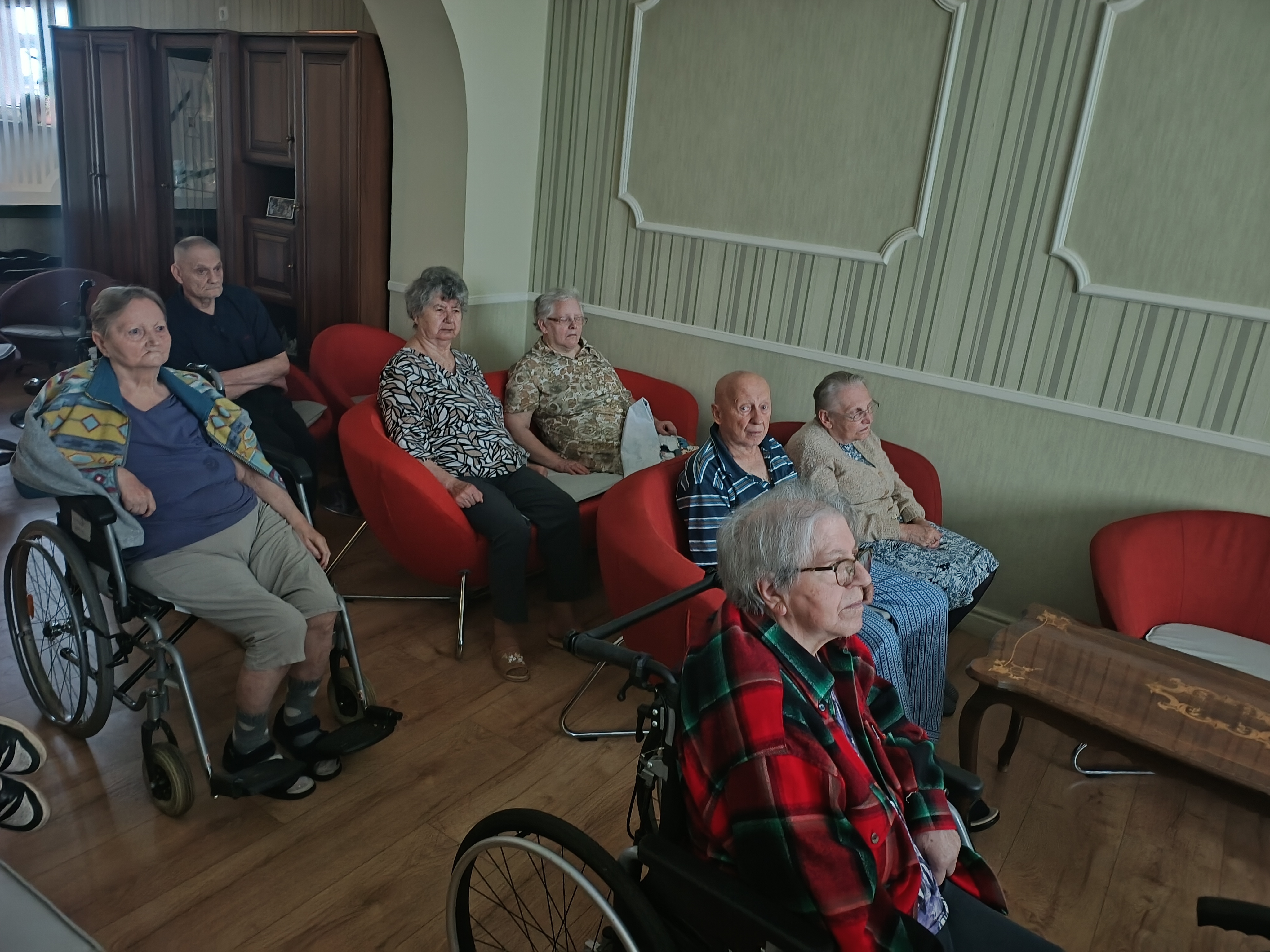 Grupa osób na kanapach i na wózkach zapatrzona w ekran