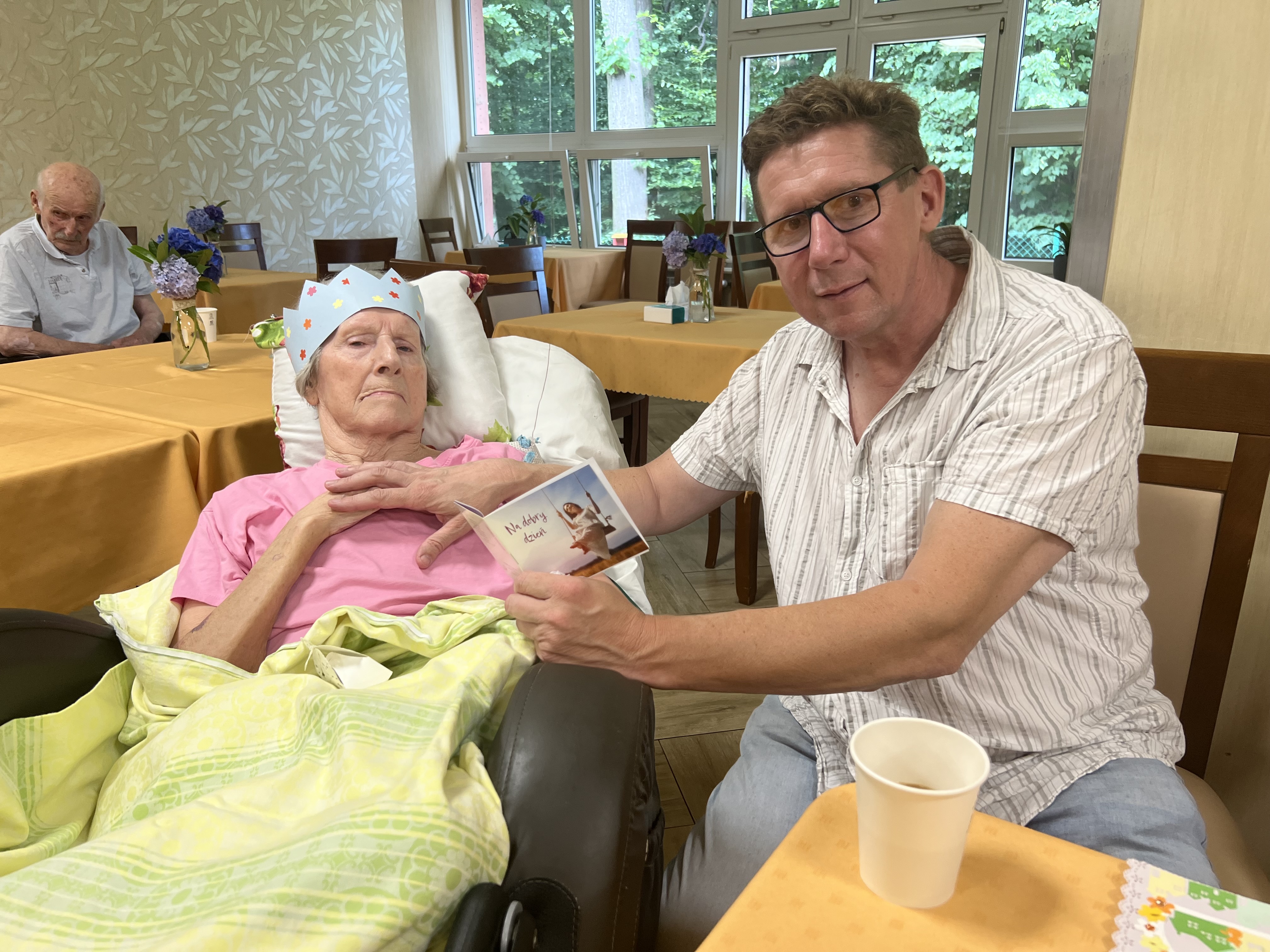 Kobieta w pozycji półleżącej w wózku geriatrycznym, na głowie ma papierową koronę, mężczyzna w okularach czyta życzenia z kartki okolicznościowej