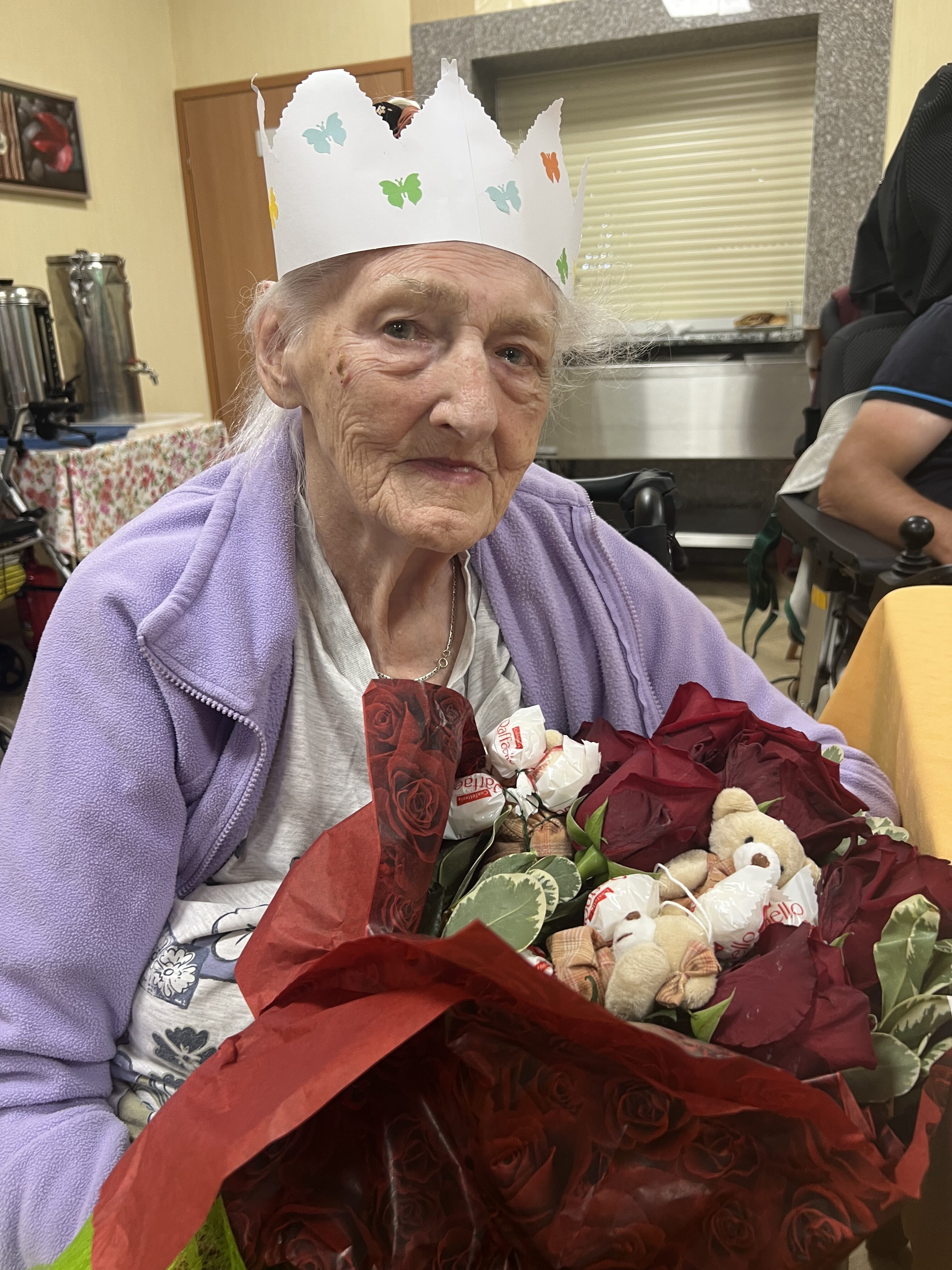 Seniorka na wózku z wielkim bukietem kwiatów, na głowie ma papierową koronę
