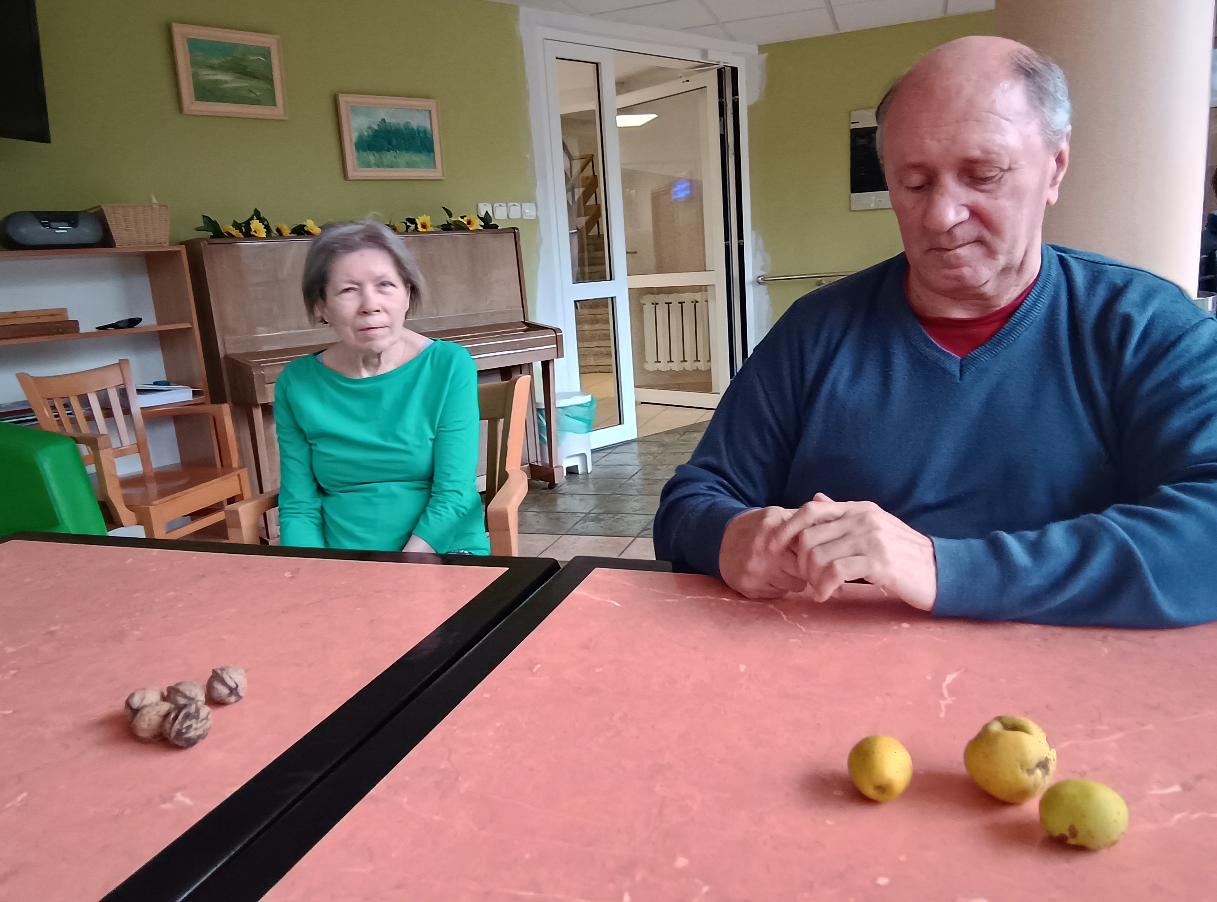 Mężczyzna i kobieta siedzą przed stołem, na stole rekwizyty do zajęć - owoce pigwowca oraz orzechy włoskie